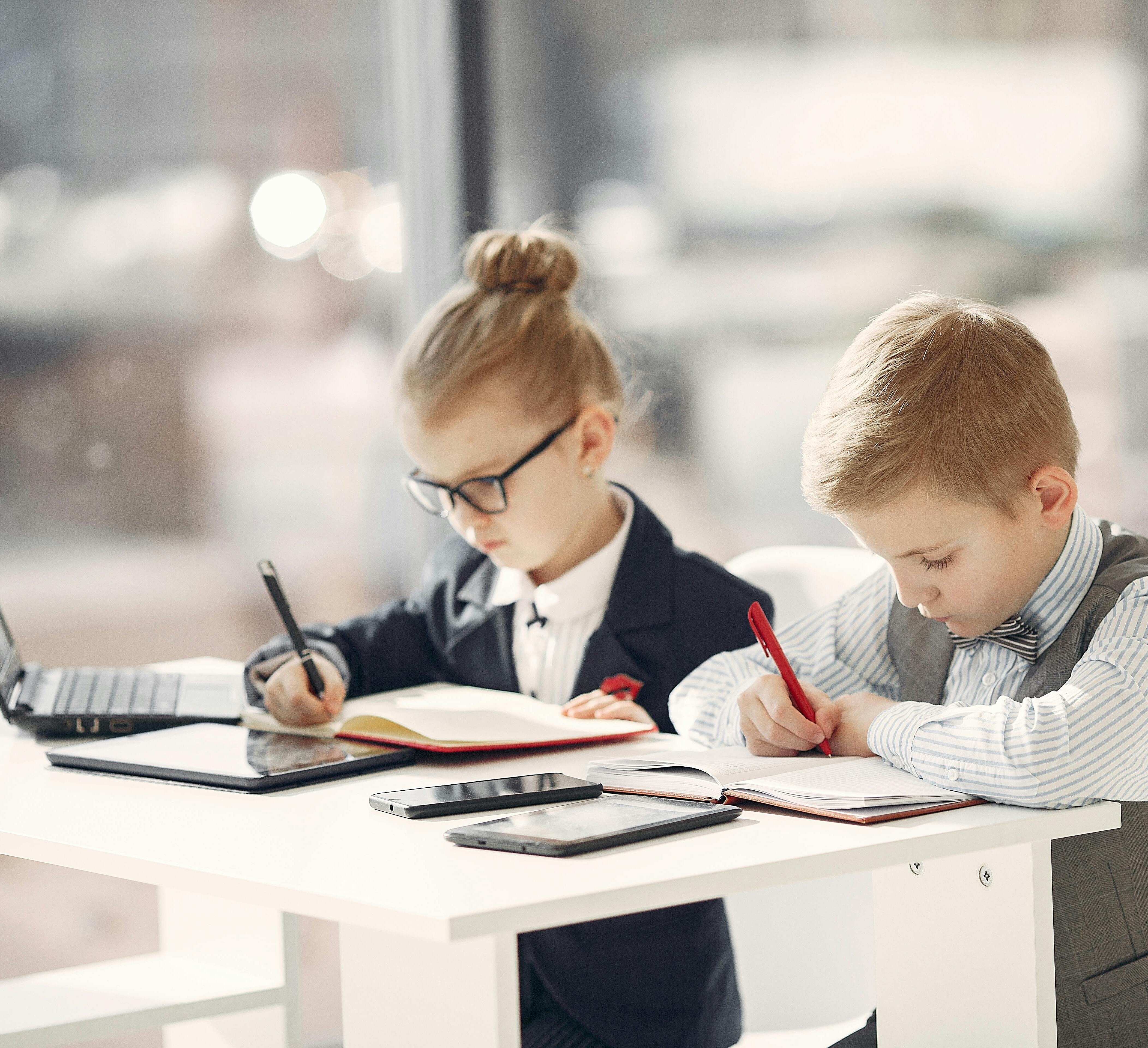 Two children writing in their books in a desk. There are various electronic devices on the desk.